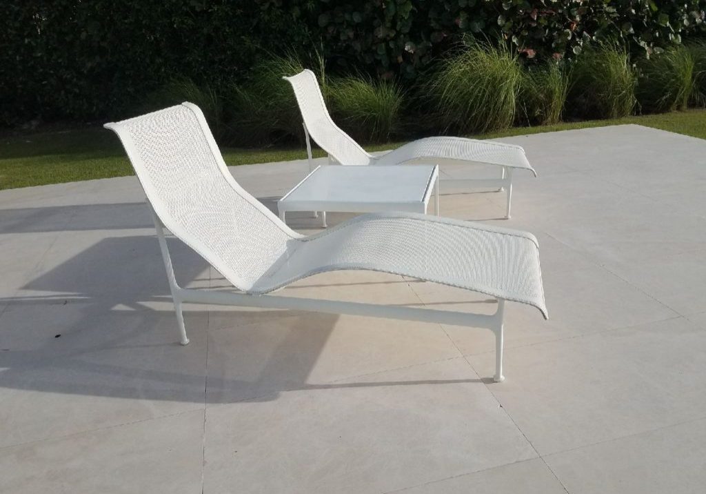 White chaise lounges on patio with greenery background.